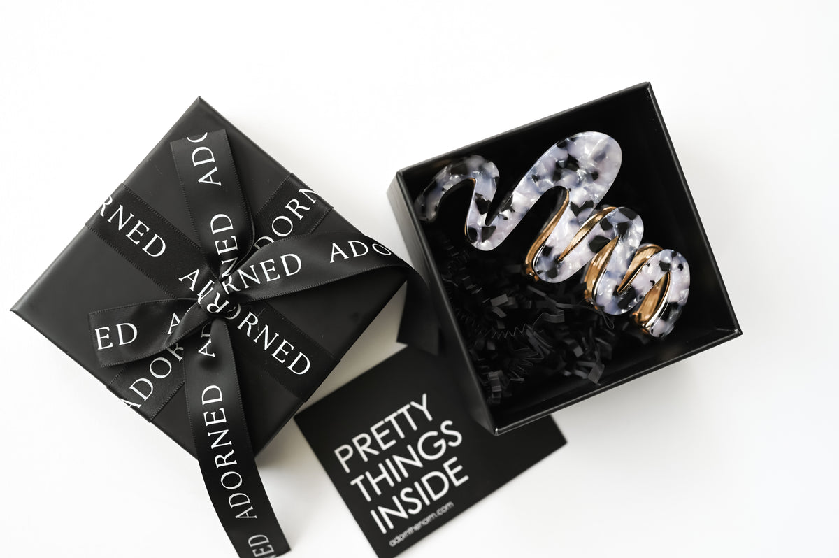 A black and gray hair claw clip with a tortoiseshell pattern and gold-tone metal accents, presented in an open black gift box  and a card reading 'PRETTY THINGS INSIDE'.