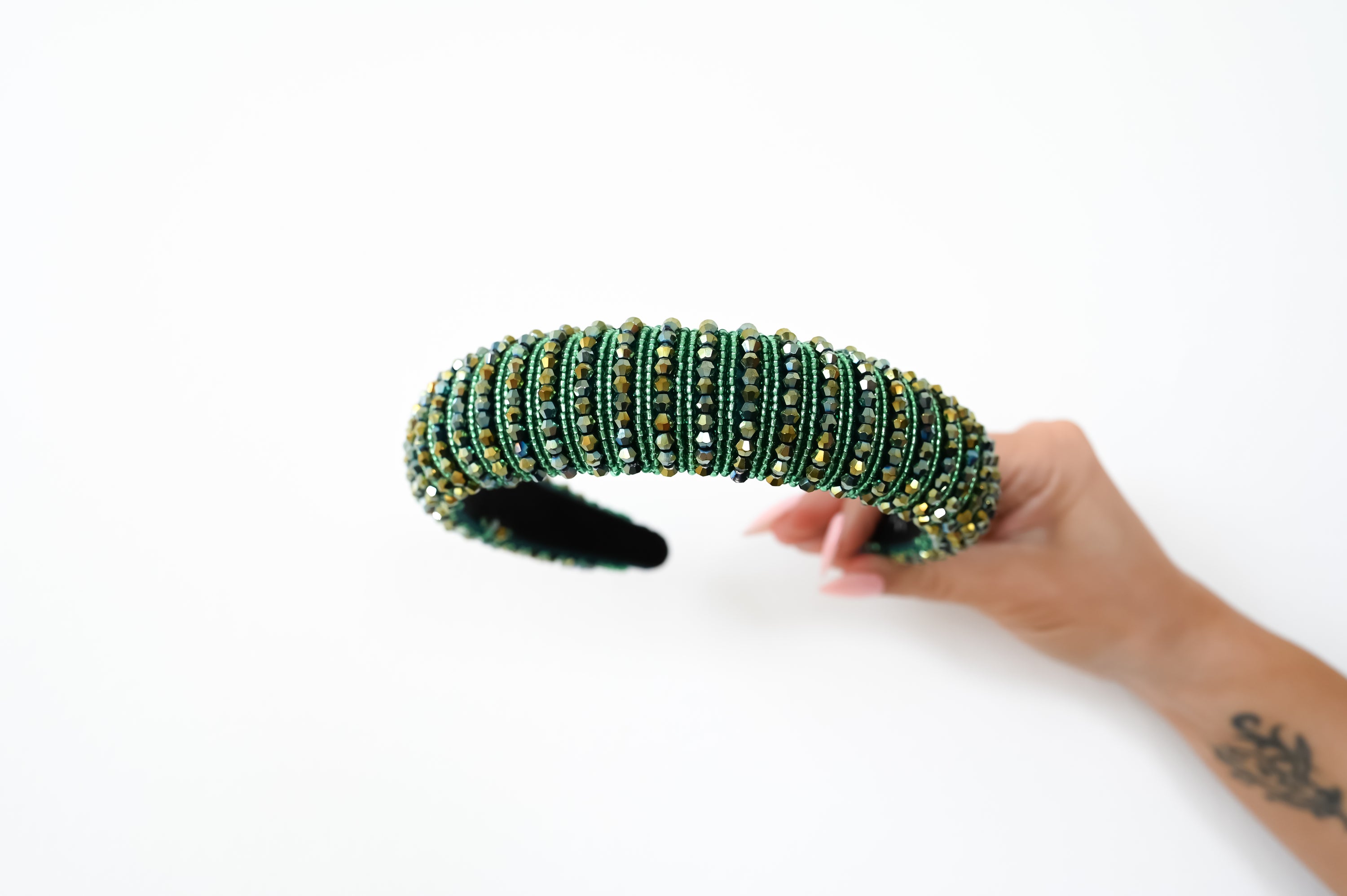 A hand holding a wide, beaded headband decorated with green, blue, and gold rhinestones, set against a plain white background.