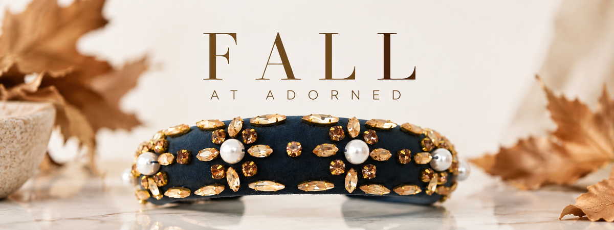 A navy blue headband embellished with gold rhinestones, marquise crystals, and white faux pearls, displayed on a marble surface with autumn leaves and the text 'FALL AT ADORNED'.